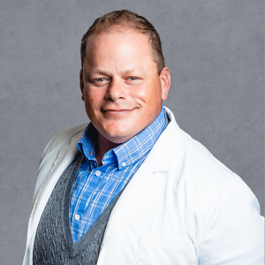 Benjamin Vandersommen, primary care provider at Optima Medical in Tatum Highlands, Phoenix, Arizona, wearing a white coat and blue shirt with a gray vest.