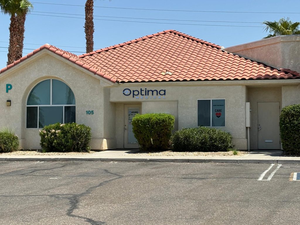 Exterior view of Optima Medical primary care clinic at 3015 AZ-95 Suite 105 in Bullhead City, Arizona, featuring tan building with red tile roof and clear front signage.