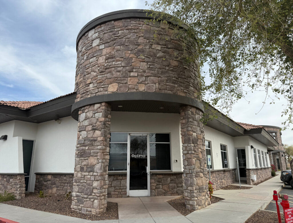 Optima Medical primary care clinic in East Surprise, Arizona, featuring a modern white building with stone façade and rounded tower entrance under a bright sky.