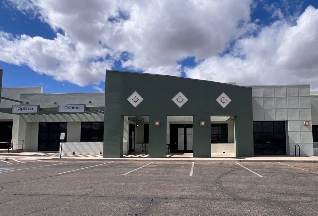 Optima Medical primary care clinic in Kingman, Arizona, featuring a modern green and white building with geometric designs and ample front parking under a partly cloudy sky.