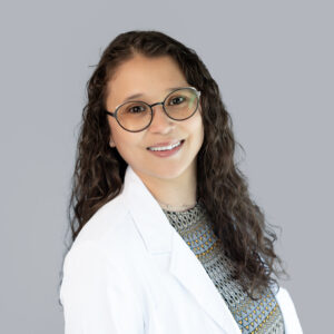 MariAngela Reynoso, primary care provider at Optima Medical in Tucson, Arizona, smiling in a white coat with glasses and curly hair.