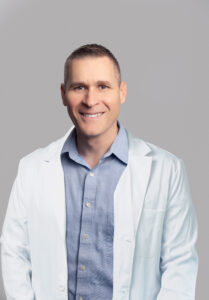 Male medical provider wearing a blue shirtand a white lab coat, smiling, posed against a light gray background.