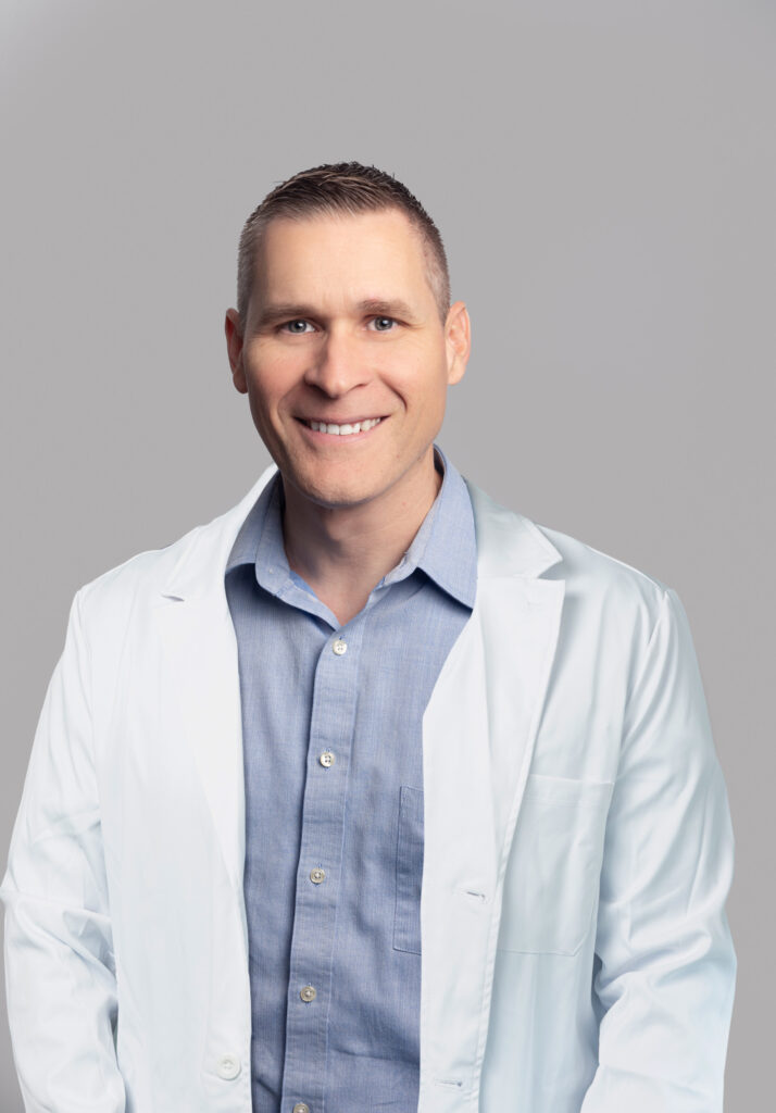 Male medical provider wearing a blue shirtand a white lab coat, smiling, posed against a light gray background.