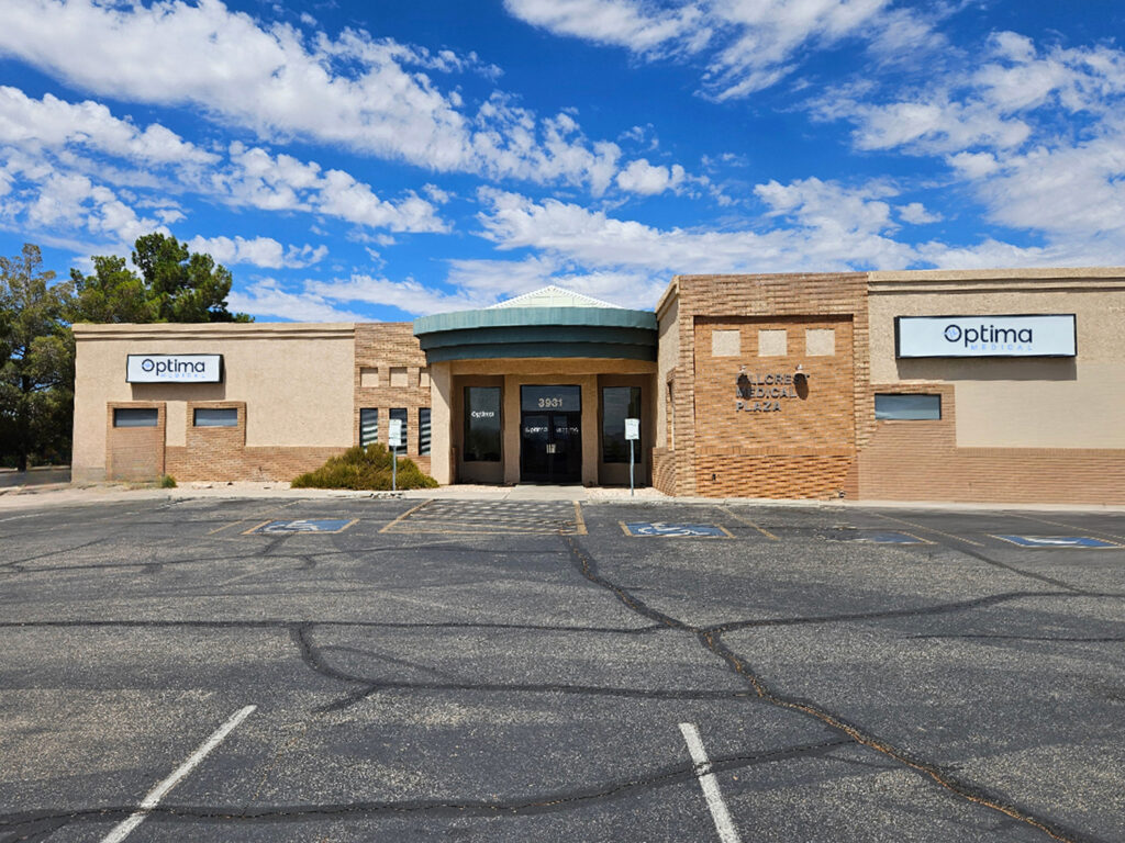 Optima Medical primary care clinic in North Kingman, Arizona, located on Stockton Hill Road with a tan brick exterior, blue roof accents, and accessible parking out front.