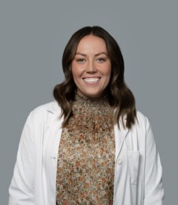Kyrie Nicolds, healthcare provider at Optima Medical in Tatum Highlands, Phoenix, Arizona, smiling in a white coat and patterned sweater.