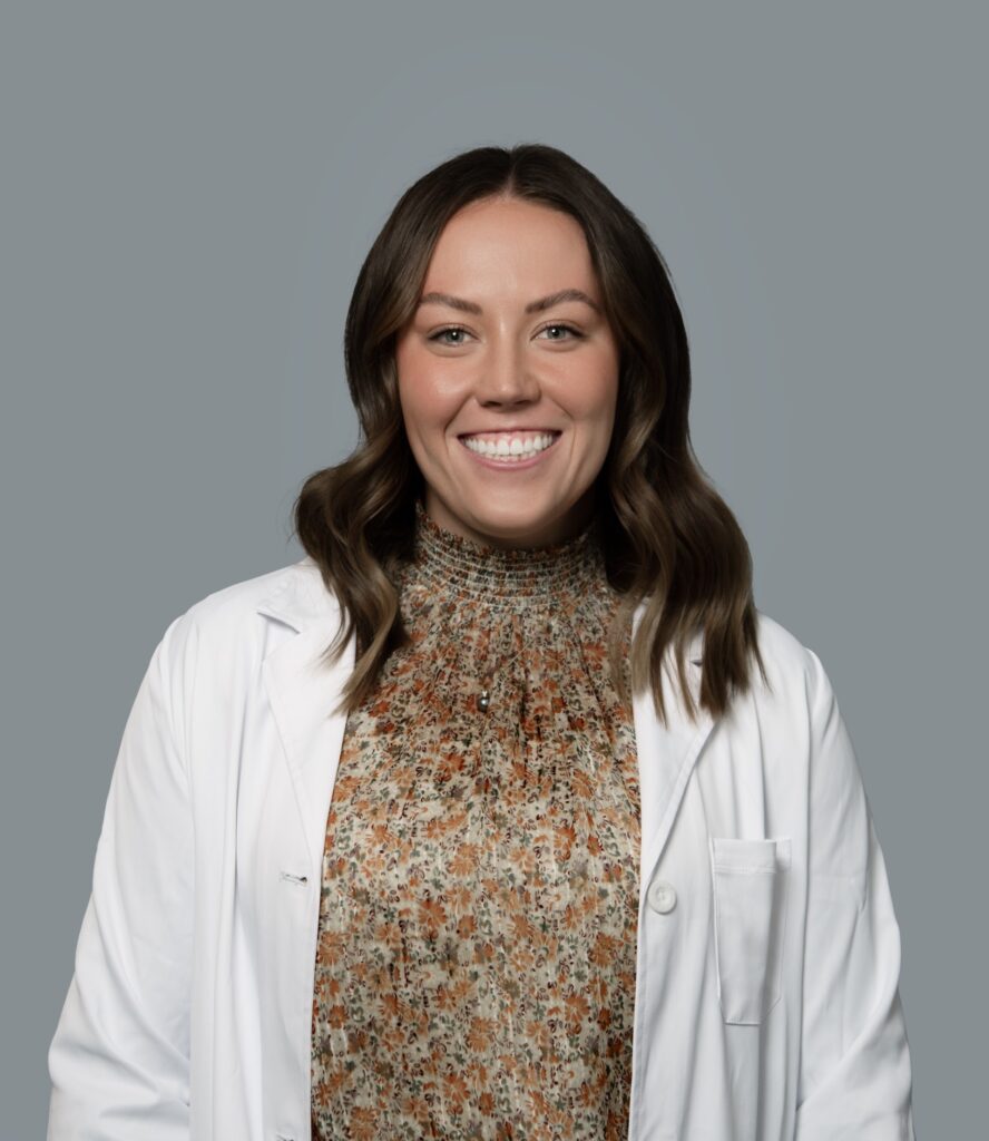 Kyrie Nicolds, healthcare provider at Optima Medical in Tatum Highlands, Phoenix, Arizona, smiling in a white coat and patterned sweater.