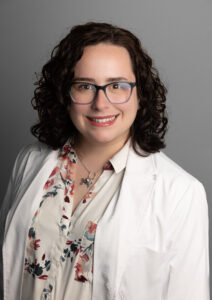 Optima Medical provider with curly dark hair and glasses, wearing a white lab coat over a floral blouse and a silver flower necklace, smiling confidently against a plain gray background.