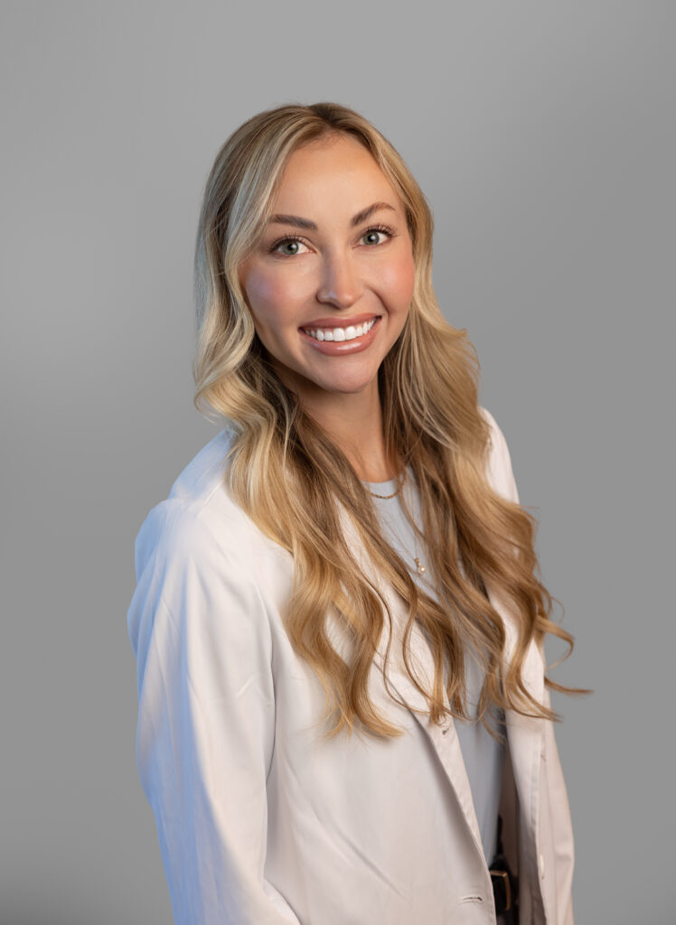 Portrait of Kodee Avery, PA-C, primary care provider at Optima Medical. Smiling professional wearing a white coat, with long blonde hair, in front of a neutral gray background.