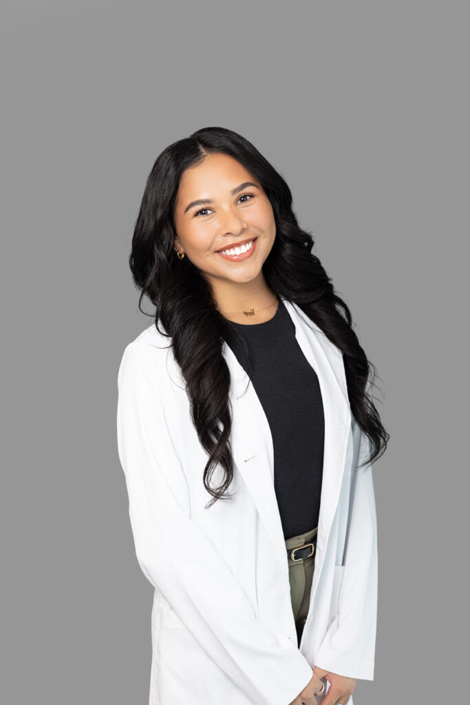 Female medical provider wearing a white lab coat, smiling, standing against a light gray background