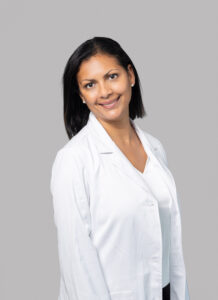 Female medical provider wearing a white lab coat, smiling, posed against a light gray background.