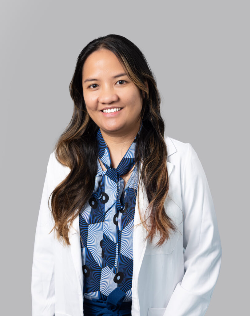 Professional headshot of a female Optima Medical provider wearing a white coat and blue patterned blouse, smiling against a neutral gray background.