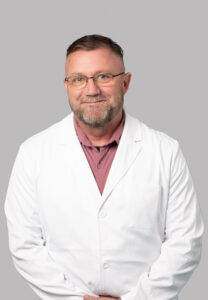 Male medical provider wearing glasses and a white lab coat, smiling, posed against a light gray background.