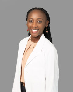 Professional headshot of a smiling Optima Medical provider wearing a white lab coat, peach blouse, and gold jewelry, with braided hair and a neutral gray background.