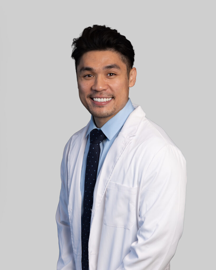 Professional headshot of a male Optima Medical provider wearing a white lab coat and light blue collared shirt, smiling against a neutral gray background.